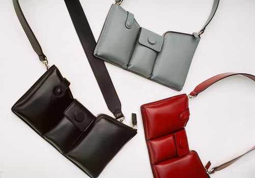 Eine junge, blonde Frau trägt ein fließendes, schwarzes One Shoulder Dress mit Cutout von KM by Lange und hält eine graue Crossbody-Bag aus genarbtem Leder in ihrer Hand.