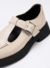 Load image into Gallery viewer, Detailaufnahme eines Mary Jane Schuhs aus cremefarbenem Leder mit dezentem Perlglanz. Das Modell verfügt über einen T-Steg, eine silberfarbene Schnalle, eine schwarze Laufsohle und schwarzes Lederfutter.
