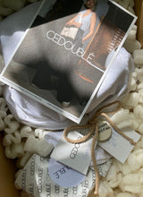 Load image into Gallery viewer, Foto von dem Inhalt eines Versandkartons von Cedoublé. Es wird gezeigt, welch aufwendige, nachhaltige Verpackung die Käufer und Käuferinnen erhalten. Auf dem Foto ist ein in einem Baumwollbeutel verpackter Artikel zu sehen, daran befestigt sind Pflegekarten und ein Tonanhänger mit liebevollem Spruch. Zusätzlich liegt eine Grußkarte in dem Paket, auf diese fällt ein sanfter Lichtstrahl.