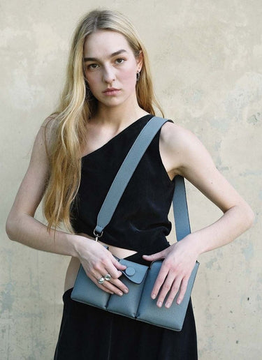 Bild eines weiblichen Models mit langen, blonden Haaren, gekleidet in einem schwarzen, langen Kleid mit seitlichem Cut-out rechts. Eine graue, genarbte Umhängetasche des Labels Cedoublé hängt ihr frontal über die linke Schulter, mit den Händen deutet die Frau ein Öffnen des mittleren Fachs mit Druckknopf an. Der Blick des Models geht direkt in die Kamera, er ist ernshaft.
