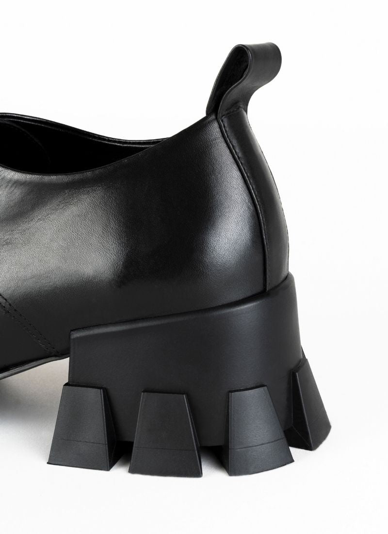 Produktbild eines schwarzen Schnürschuhs mit Karree Leisten und Anziehlasche. Im Fokus des Bildes steht der markante, skulpturale Absatz mit breiter Auftrittsfläche und Profil. Der Absatz ist ca. 5,5 cm hoch.  