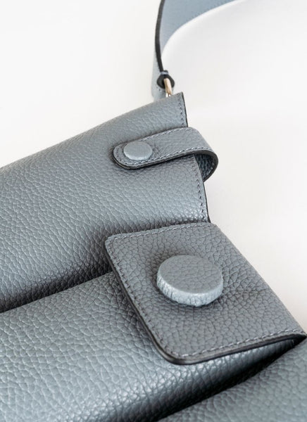 Detailfoto einer Crossbody-Bag aus grauem, genarbtem Leder mit drei voneinander abgetrennten Fächern und einem farblich passenden, breiten Gurt aus Leder mit Karabinerhaken. Diese Handtasche ist genderfluid, angefertigt wurde sie von dem Newcomer-Label Cedoublé.