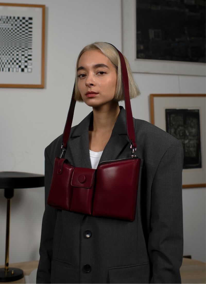 Eine weinrote Crossbody-Bag aus weich gepolstertem Glattleder mit drei voneinander abgetrennten Fächern hängt um den Kopf eines weiblichen Models mit kinnlangen, blonden Haaren. Das Model trägt einen grauen Oversize-Blazer mit Schulterpolstern.
