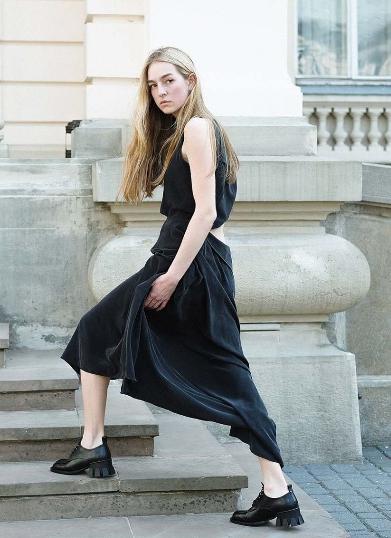 Bild eines Models mit langen, blonden Haaren, welches mit ernstem Blick die Treppe eines historischen Gebäudes hinauf schreitet. Das schwarze, lange Kleid verfügt über einen Cut-out am Rücken. An den Füßen trägt die Dame extravagante Leder-Schnürschuhe mit einem ausdrucksstarken Absatz. Der Kontrast zwischen femininer Kleidung und klobigeren Schuhen ist hier sehr schön gelungen.