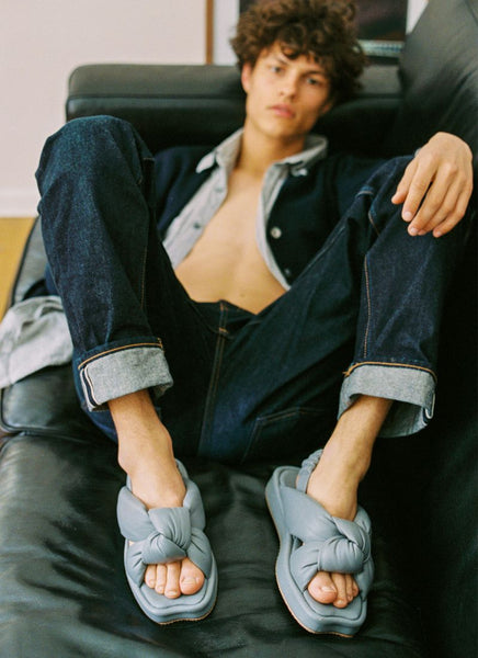 Foto eines männlichen Models mit lockigem, kurzem Haar. Er liegt lässig auf einer schwarzen Ledercouch, trägt eine umgekrempelte Jeans und ein offenes Oberteil, sodass seine trainierte Brust entblößt ist. An den Füßen trägt er graue puffy Sandalen  aus Leder des Herstellers Cedoublé.