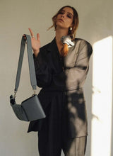 Lade das Bild in den Galerie-Viewer, Bild eines weiblichen Models, welches in einen oversized, grauen Herrenanzug gekleidet ist. Ihre langen, braunen Haare sind mit einem grauen Cord-Scrunchie zu einem seitlichen, lockeren Zopf gebunden, sie trägt Silberschmuck. Den linken Arm hält sie vor der Brust verschränkt, der rechte Arm ist angewinkelt. An diesem baumelt locker eine hellgraue, hochwertige Designertasche. Diese ist schmal in der Tiefe und verfügt über Karabiner, sodass der Gurt ausgetauscht werden kann.
