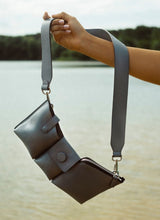 Lade das Bild in den Galerie-Viewer, Die sehr gepflegte Hand einer jungen Frau hält eine graue Crossbody Bag am ausgestreckten Arm in die Höhe. Im Hintergrund befindet sich ein See und weiter entfernt sind Bäume zu sehen. Bei der Tasche handelt es sich um das graue Polly Modell des Labels Cedouble. Eine sehr kompakte Variante, die sich wunderbar an den Körper anschmiegt und ganzjährig getragen werden kann.
