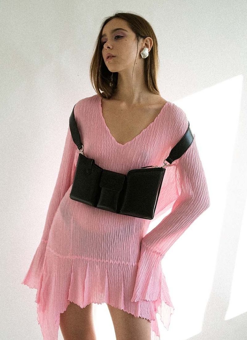 Foto eines weiblichen Models mit langen braunen Haaren. Sie trägt ein leicht transparentes, hell pinkfarbenes Kleid mit gezackten Säumen, eine schwarze Handtasche aus Leder ist künstlerisch um beide Schultern gehängt, der Taschenkörper zeigt nach vorne und baumelt etwa auf Unterbrusthöhe der Frau. Das Model hat ihren Blick zur Seite gerichtet, sie steht vor einem weißen Hintergrund. Die Tasche ist von der Marke Cedoublé.